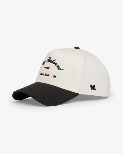 Van Libra Bone/Carbon (cream and dark gray) structured cap with bone colored crown and carbon colored brim. Features black embroidered text “Stay Balanced” with red abstract triangle graphic on the front, black “1986” and “VAN LIBRA” details, and “VL” stitched on the side, finished with an adjustable carbon colored snapback closure at the back.