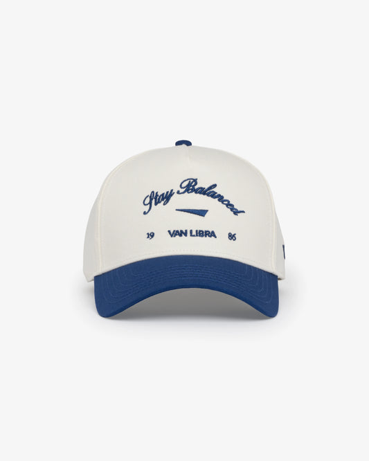Van Libra Bone/Indigo (cream and blue) structured cap with bone colored crown and indigo colored brim. Features blue embroidered text “Stay Balanced” with abstract triangle graphic on the front, “1986” and “VAN LIBRA” details, and “VL” stitched on the side, finished with an adjustable indigo snapback closure at the back.