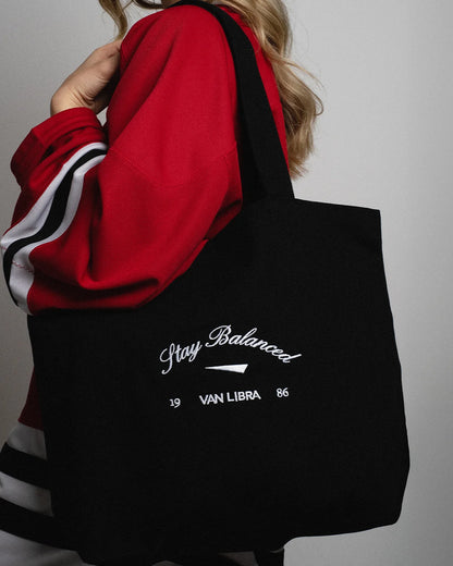 Van Libra Blackwood (black) canvas tote bag with white embroidery. Features “Stay Balanced” text above an abstract triangle graphic, with “VAN LIBRA” and the numbers “19” and “86” stitched below.