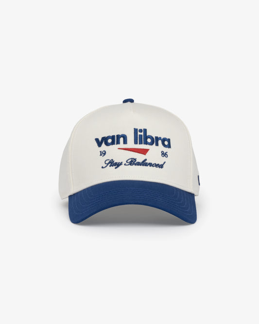 Van Libra Bone/Indigo (cream and blue) structured cap with bone colored crown and indigo colored brim. Features blue embroidered text “van libra” with red abstract triangle graphic on the front, blue “1986” and “Stay Balanced” lettering on the front, plus “VL” stitched on the side, finished with an adjustable indigo snapback closure at the back.