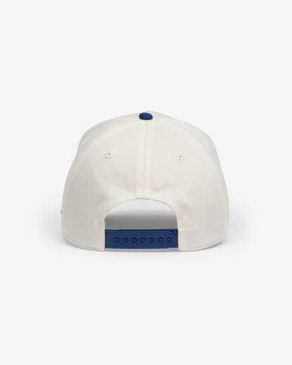 Van Libra Bone/Indigo (cream and blue) structured cap with bone colored crown and indigo colored brim. Features blue embroidered text “van libra” with red abstract triangle graphic on the front, blue “1986” and “Stay Balanced” lettering on the front, plus “VL” stitched on the side, finished with an adjustable indigo snapback closure at the back.